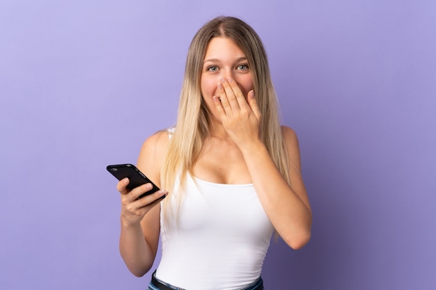 Young blonde woman using mobile phone isolated on purple with surprise facial expression