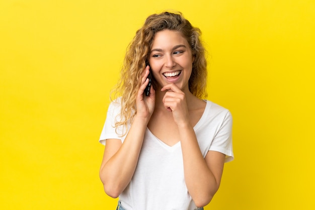 Young blonde woman using mobile phone isolated on yellow background looking to the side and smiling