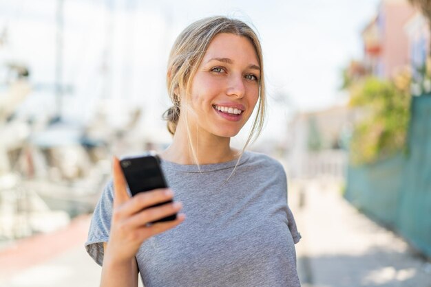 幸せな表情で屋外で携帯電話を使用する若いブロンドの女性