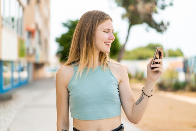 幸せな表情で屋外で携帯電話を使用する若いブロンドの女性