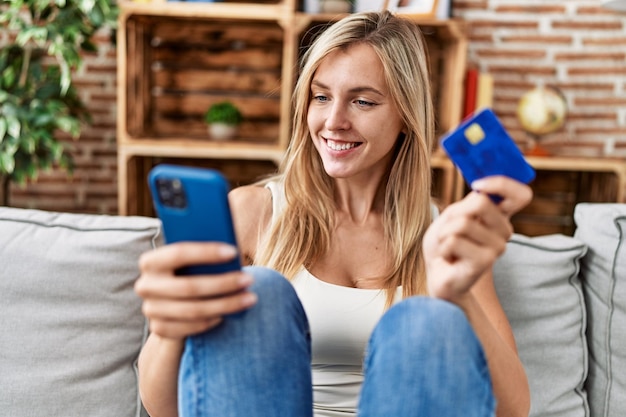 Young blonde woman using smartphone and credit card sitting on sofa at home