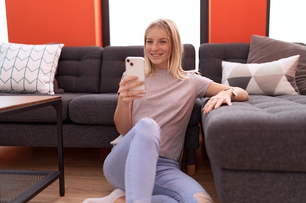 Young blonde woman using smartphone sitting on floor at home