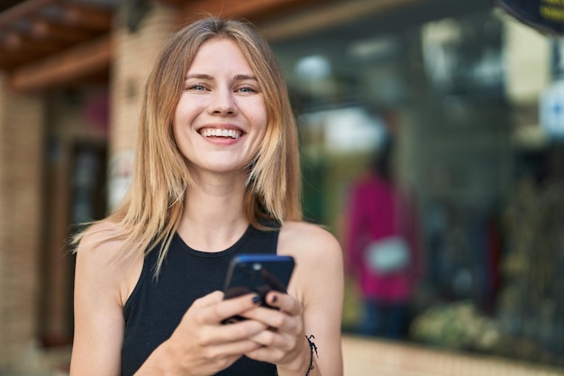 Young blonde woman using smartphone smiling at street