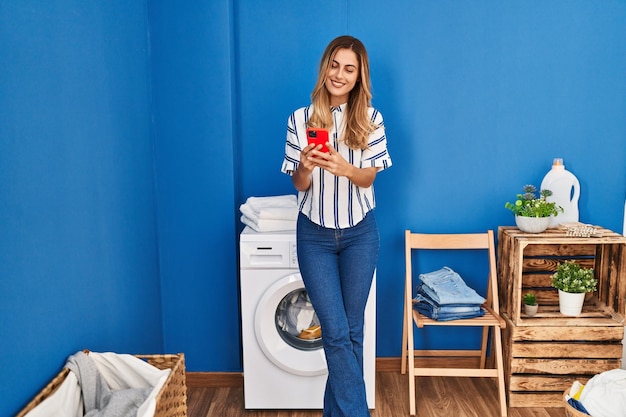 Young blonde woman using smartphone waiting for washing machine at laundry room