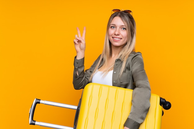 Young blonde woman in vacation with travel suitcase and making victory gesture