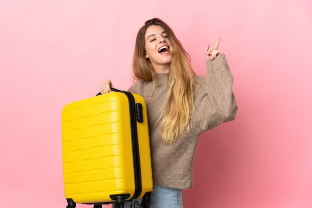 Young blonde woman over in vacation with travel suitcase and pointing up