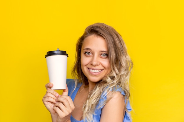 Una giovane donna bionda con i capelli ondulati che tiene in mano una tazza di caffè o tè di carta bianca con spazio vuoto per la copia