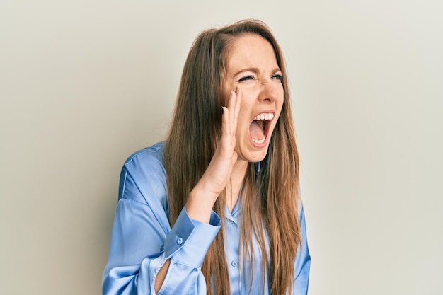 Young blonde woman wearing casual blue shirt shouting and screaming loud to side with hand on mouth communication concept