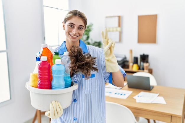 Young blonde woman wearing cleaner uniform holding cleaning products smiling with happy face winking at the camera doing victory sign with fingers. number two.