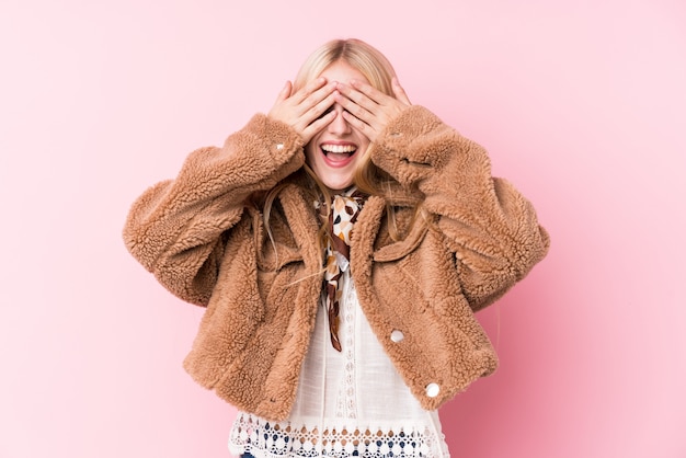 Young blonde woman wearing a coat against a pink wall covers eyes with hands, smiles broadly waiting for a surprise.