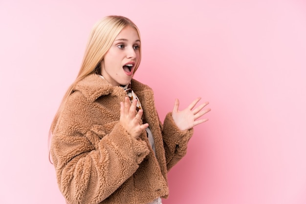 Young blonde woman wearing a coat against a pink wall shouts loud, keeps eyes opened and hands tense.