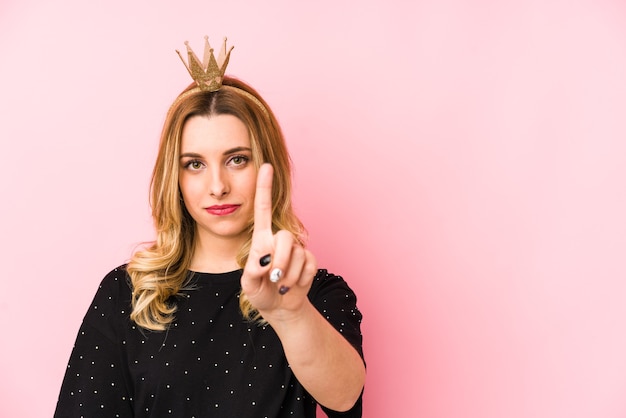 Young blonde woman wearing a crown isolated showing number one with finger.