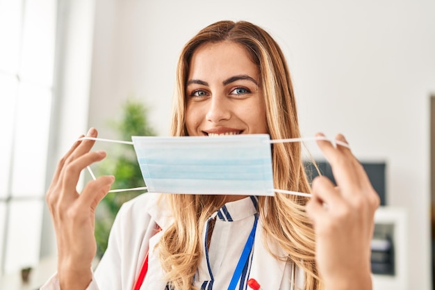 Young blonde woman wearing doctor uniform holding medical mask at clinic