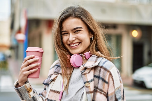 Young blonde woman wearing headphones holding coffee at street