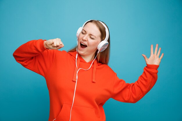 Young blonde woman wearing headphones raising hand pretend holding microphone singing with closed eyes 