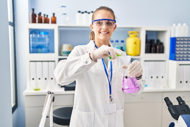 Young blonde woman wearing scientist uniform pouring liquid on test tube at laboratory