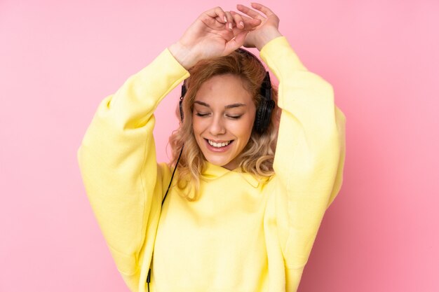 Young blonde woman wearing a sweatshirt isolated on pink wall listening music and dancing