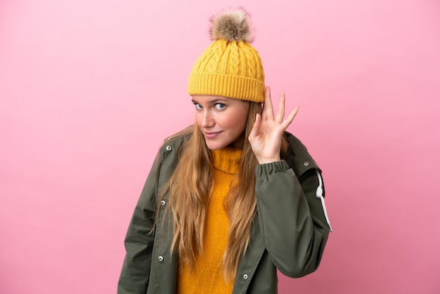 Photo young blonde woman wearing winter jacket isolated on pink background listening to something by putting hand on the ear