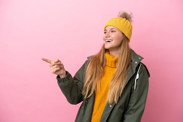 Young blonde woman wearing winter jacket isolated on pink background pointing finger to the side and presenting a product