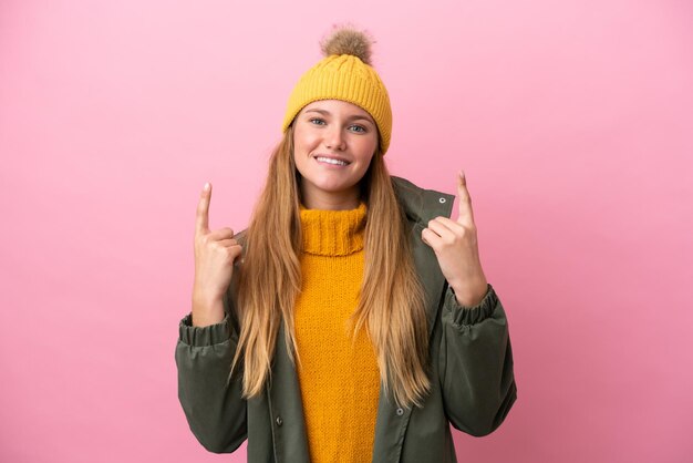 Young blonde woman wearing winter jacket isolated on pink background pointing up a great idea