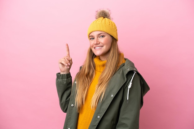 Young blonde woman wearing winter jacket isolated on pink background showing and lifting a finger in sign of the best