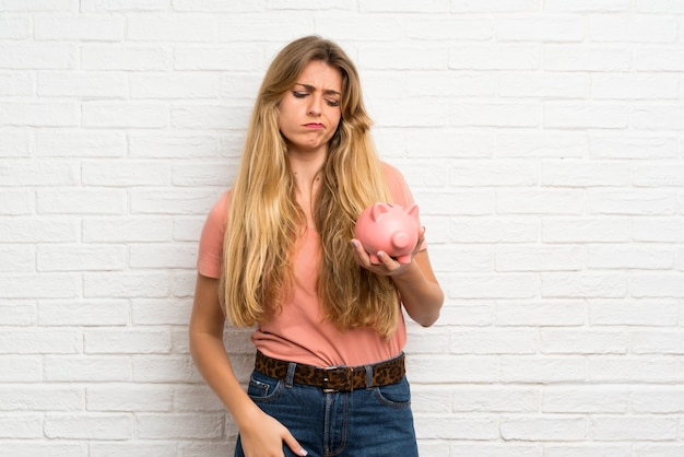 Young blonde woman over white brick wall holding a big piggybank