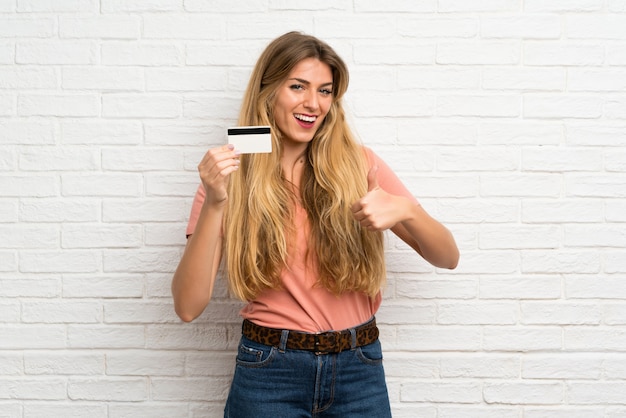 Young blonde woman over white brick wall holding a credit card