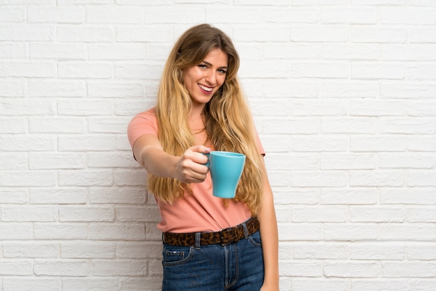 Giovane donna bionda sopra il muro di mattoni bianco che tiene tazza di caffè calda