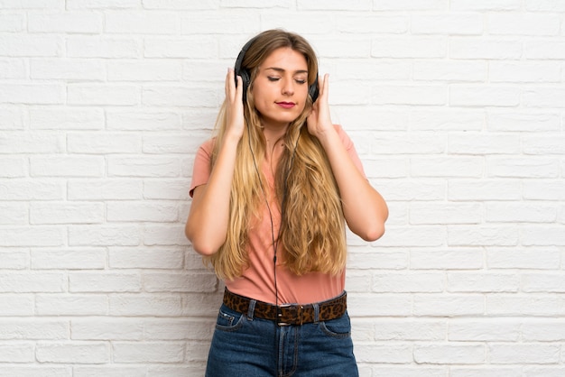 Молодая белокурая женщина над белой кирпичной стеной слушая к музыке с наушниками