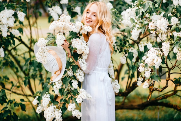 Young  blonde woman in white dress posing in lilac bushes,