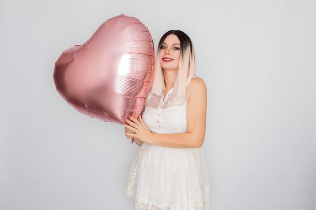 Young blonde woman in white lace dress holding big pink heart balloon in her hands over white background