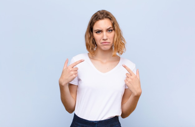 Young blonde woman with a bad attitude looking proud and aggressive, pointing upwards or making fun sign with hands against flat color wall