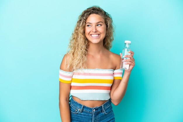 Young blonde woman with a bottle of water isolated on blue background looking side