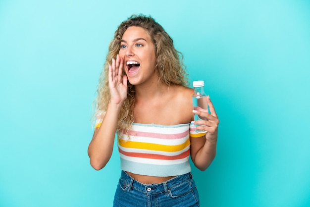 Young blonde woman with a bottle of water isolated on blue background shouting with mouth wide open to the side