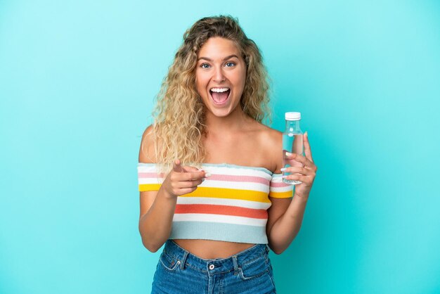 Young blonde woman with a bottle of water isolated on blue background surprised and pointing front