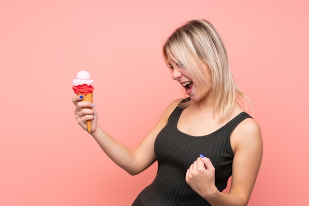 勝利を祝う分離のピンクの壁の上のコルネットアイスクリームを持つ若いブロンドの女性