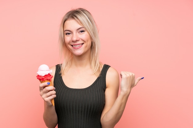 Young blonde woman with a cornet ice cream over isolated pink wall pointing to the side to present a product