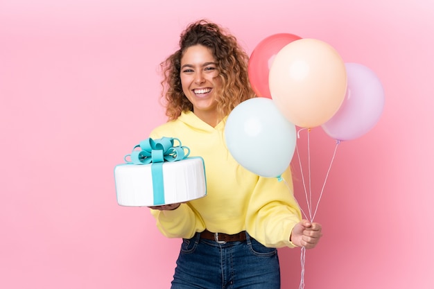 Young blonde woman with curly hair catching many balloons and holding a big cake isolated on pink