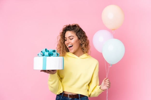 Young blonde woman with curly hair catching many balloons and holding a big cake on pink wall