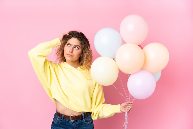 Young blonde woman with curly hair catching many balloons isolated on pink having doubts and with confuse face expression