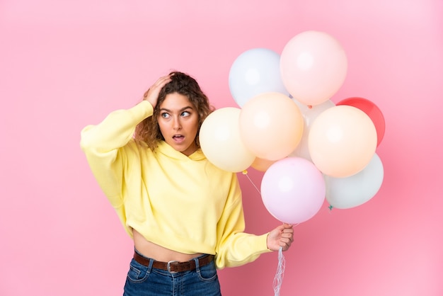 Young blonde woman with curly hair catching many balloons isolated on pink wall with surprise facial expression