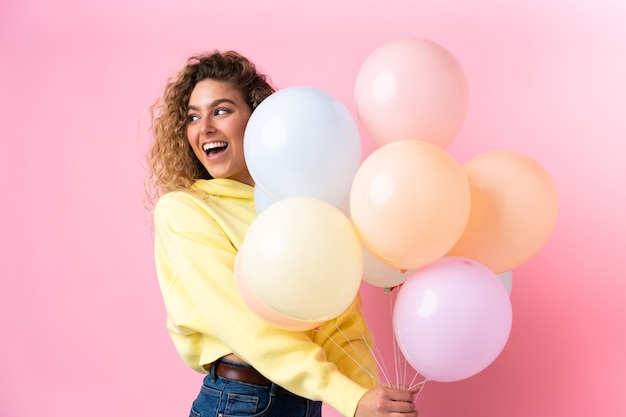 Young blonde woman with curly hair catching many balloons isolated on pink wall