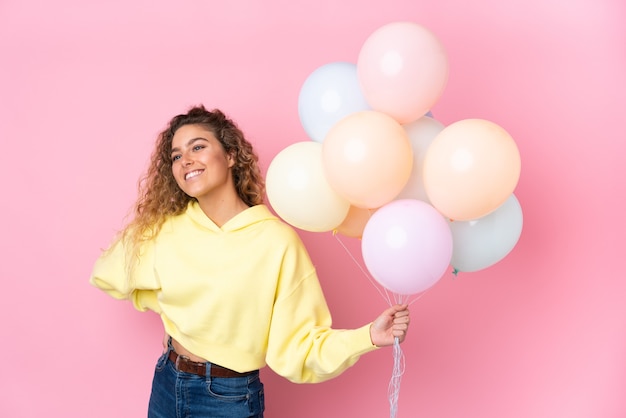 Young blonde woman with curly hair catching many balloons isolated on pink