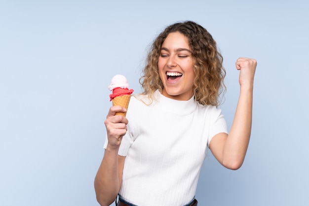 Young blonde woman with curly hair holding a cornet ice cream isolated on blue celebrating a victory