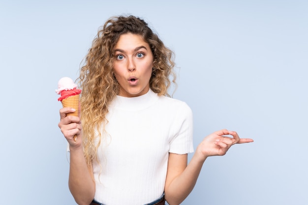 Young blonde woman with curly hair holding a cornet ice cream isolated on blue surprised and pointing side