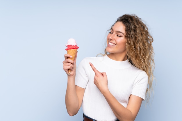 Young blonde woman with curly hair holding a cornet ice cream isolated on blue wall and pointing it