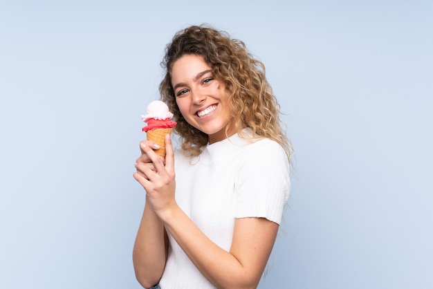 Young blonde woman with curly hair holding a cornet ice cream isolated on blue wall