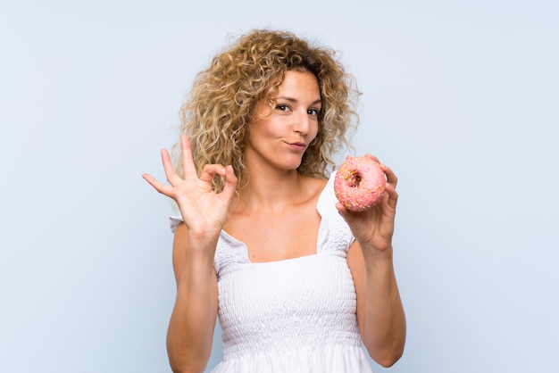 Молодая белокурая женщина с вьющимися волосами держит пончик над синей стеной, показывая знак ОК с пальцами