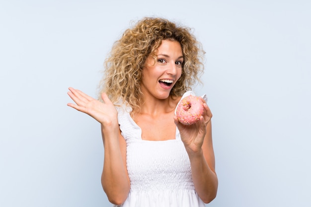 Young blonde woman with curly hair holding a donut over isolated blue wall with shocked facial expression