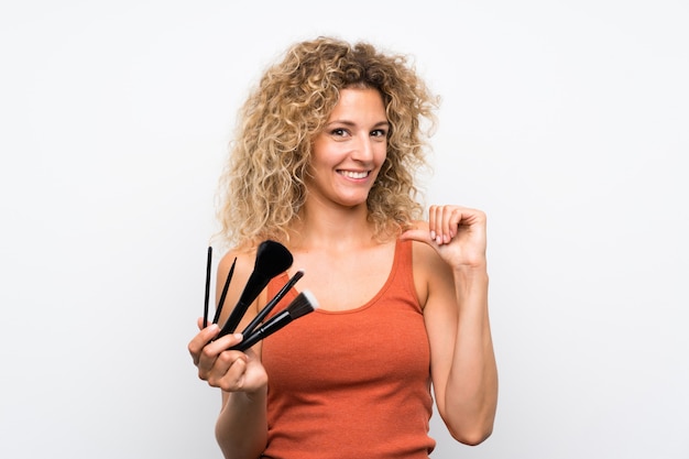 Young blonde woman with curly hair holding a lot of makeup brush proud and self satisfied